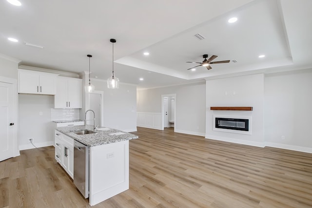 kitchen with sink, light stone counters, a raised ceiling, an island with sink, and white cabinets