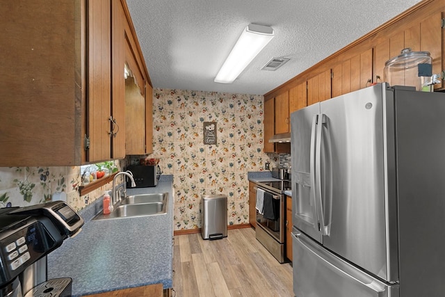 kitchen with a textured ceiling, stainless steel appliances, a sink, brown cabinets, and wallpapered walls