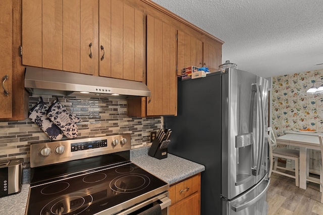 kitchen with a textured ceiling, under cabinet range hood, stainless steel appliances, light wood-type flooring, and brown cabinetry