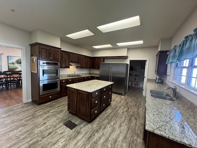 kitchen with dark brown cabinetry, under cabinet range hood, stainless steel appliances, wood finished floors, and a sink