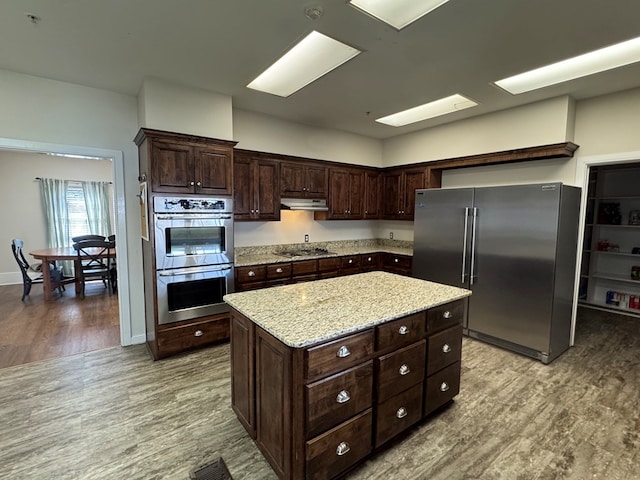 kitchen with dark brown cabinetry, a kitchen island, light stone countertops, stainless steel appliances, and light wood-style floors