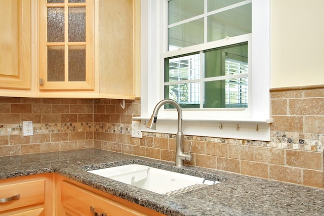 kitchen with a sink, backsplash, dark stone counters, light brown cabinetry, and glass insert cabinets