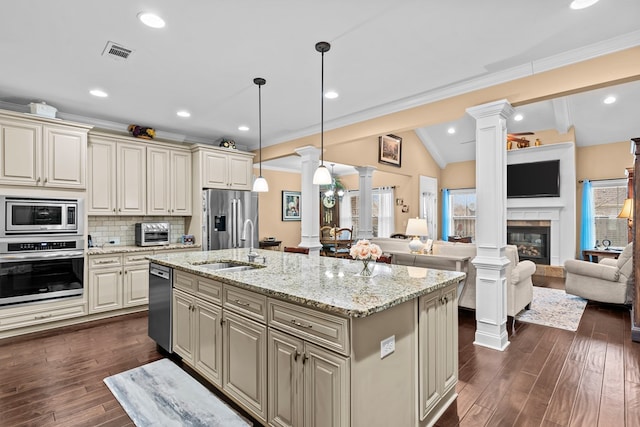 kitchen with cream cabinets, backsplash, hanging light fixtures, a kitchen island with sink, and appliances with stainless steel finishes