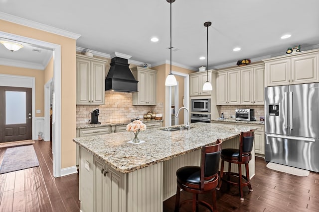 kitchen featuring stainless steel appliances, sink, cream cabinets, custom range hood, and an island with sink