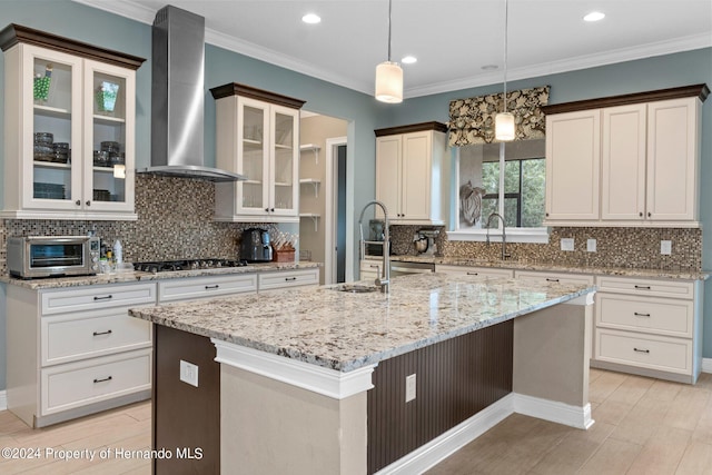 kitchen featuring stainless steel gas stovetop, hanging light fixtures, an island with sink, crown molding, and wall chimney range hood