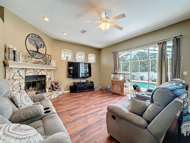 living room featuring a stone fireplace, wood-type flooring, a textured ceiling, and ceiling fan