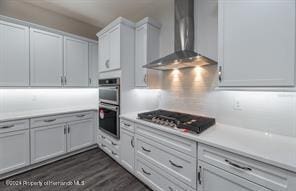 kitchen with white cabinetry, wall chimney exhaust hood, gas cooktop, double oven, and dark hardwood / wood-style flooring