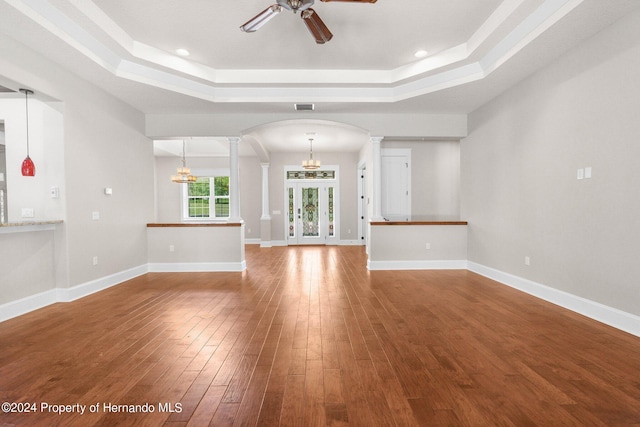 unfurnished living room with ceiling fan with notable chandelier, a tray ceiling, hardwood / wood-style flooring, and decorative columns