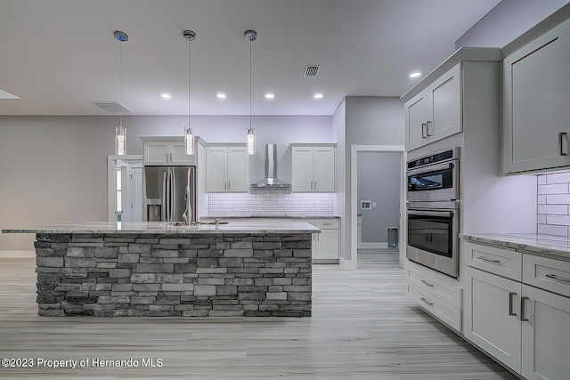 kitchen with appliances with stainless steel finishes, tasteful backsplash, wall chimney exhaust hood, decorative light fixtures, and a kitchen island with sink