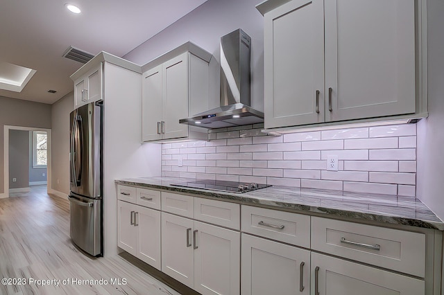 kitchen with black electric cooktop, wall chimney range hood, stone countertops, light hardwood / wood-style flooring, and stainless steel fridge