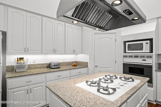 kitchen featuring light stone counters, range hood, white cabinetry, a kitchen island, and white appliances