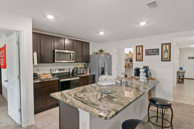 kitchen featuring appliances with stainless steel finishes, dark brown cabinetry, light stone countertops, an island with sink, and a breakfast bar area