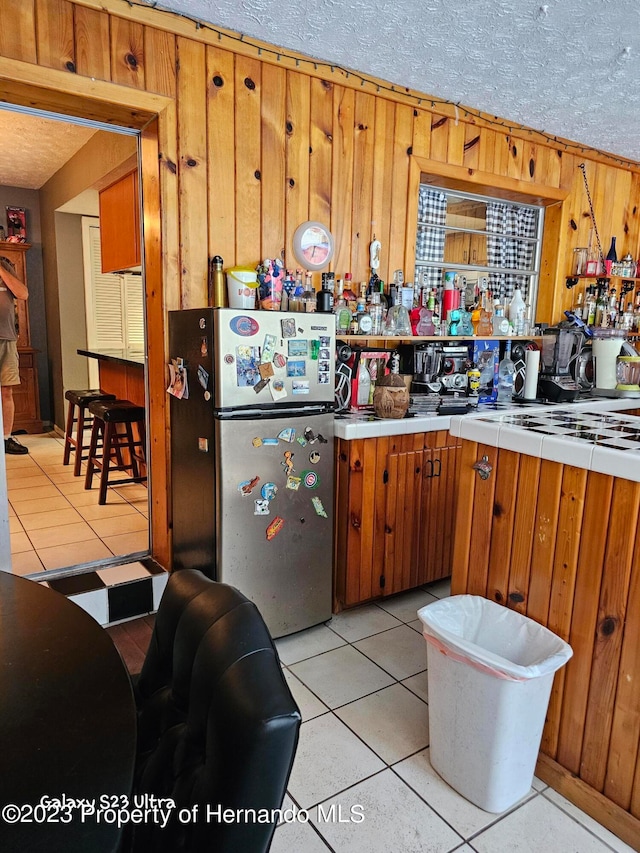 kitchen with wooden walls, a textured ceiling, light tile patterned floors, and stainless steel fridge