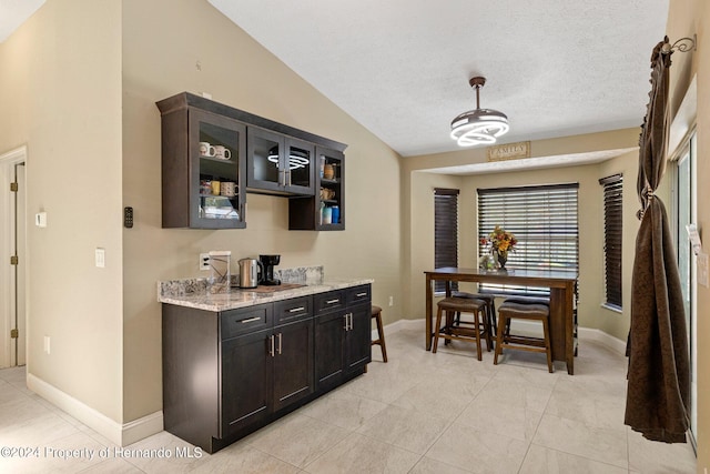 interior space featuring pendant lighting, vaulted ceiling, a textured ceiling, light stone countertops, and dark brown cabinets