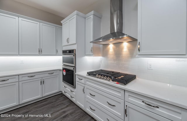 kitchen with stainless steel appliances, dark wood-type flooring, wall chimney range hood, backsplash, and white cabinetry