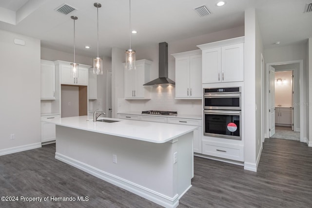 kitchen featuring white cabinetry, wall chimney range hood, appliances with stainless steel finishes, dark hardwood / wood-style flooring, and a kitchen island with sink