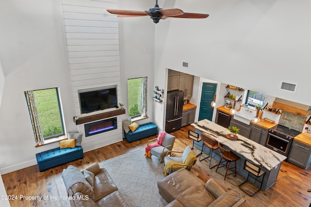 living room with ceiling fan, a high ceiling, wood-type flooring, and a large fireplace