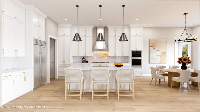 kitchen featuring stainless steel appliances, hanging light fixtures, an island with sink, wall chimney range hood, and light wood-type flooring