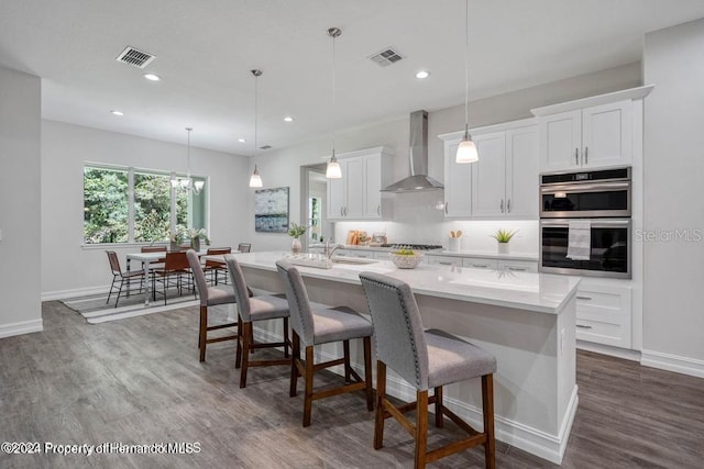 kitchen with white cabinetry, appliances with stainless steel finishes, decorative light fixtures, an island with sink, and wall chimney exhaust hood