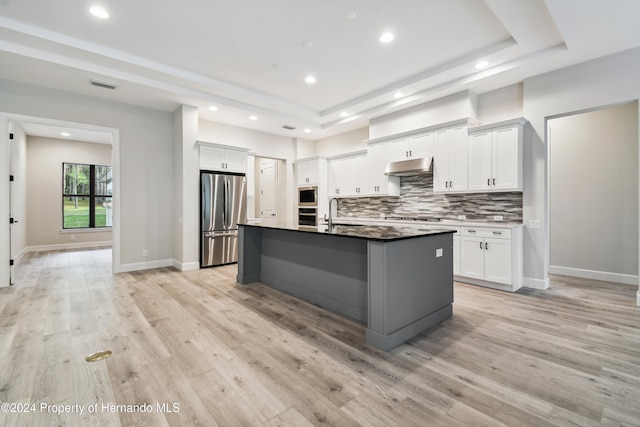 kitchen featuring stainless steel appliances, a tray ceiling, a center island with sink, white cabinets, and light hardwood / wood-style floors