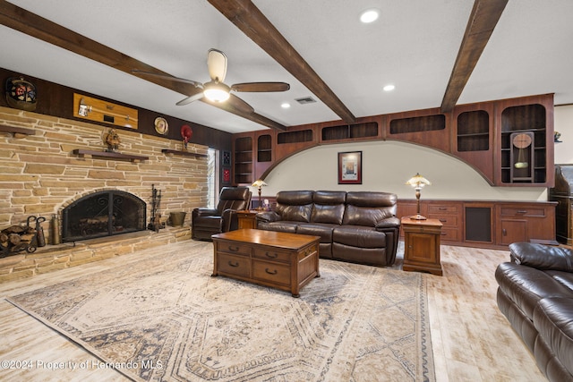 living room featuring beamed ceiling, light hardwood / wood-style flooring, ceiling fan, and a stone fireplace