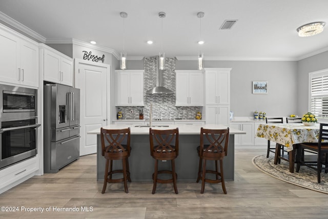 kitchen featuring stainless steel appliances, a kitchen island with sink, wall chimney range hood, pendant lighting, and white cabinetry