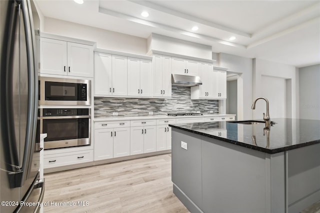 kitchen with a center island with sink, dark stone countertops, light wood-type flooring, appliances with stainless steel finishes, and white cabinetry