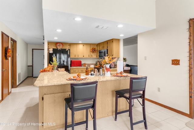 kitchen featuring light stone countertops, light brown cabinets, black refrigerator, a breakfast bar, and light tile patterned flooring