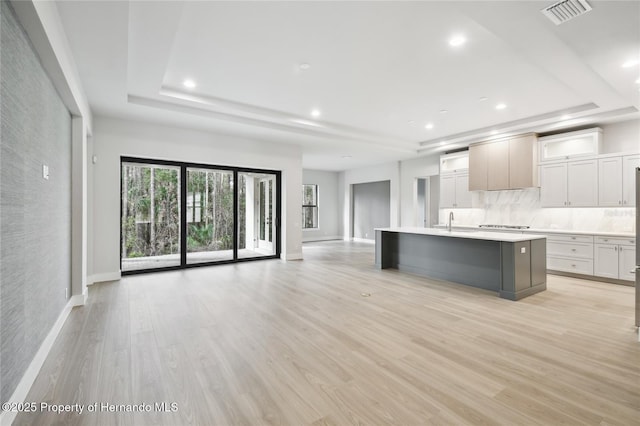 kitchen with a center island with sink, decorative backsplash, a raised ceiling, and light wood-type flooring