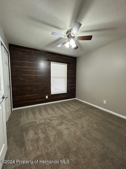 unfurnished bedroom with a textured ceiling, dark carpet, ceiling fan, and wood walls