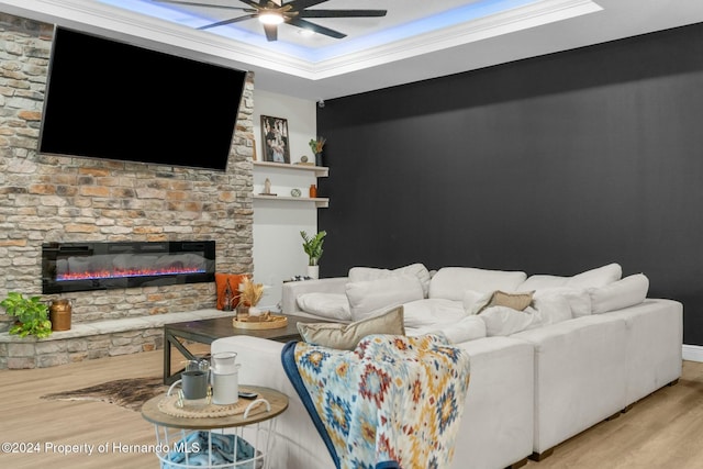 living room with light wood-type flooring, a raised ceiling, ceiling fan, crown molding, and a stone fireplace