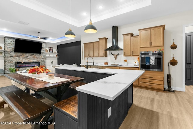 kitchen with pendant lighting, stainless steel oven, a kitchen island with sink, a raised ceiling, and wall chimney exhaust hood