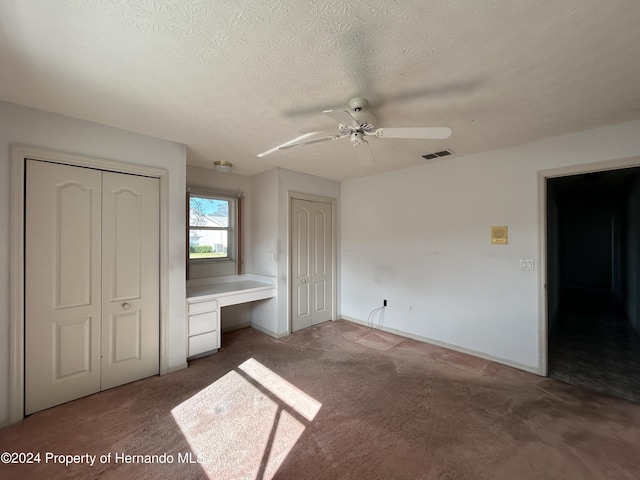 unfurnished bedroom with dark carpet, ceiling fan, built in desk, a textured ceiling, and multiple closets