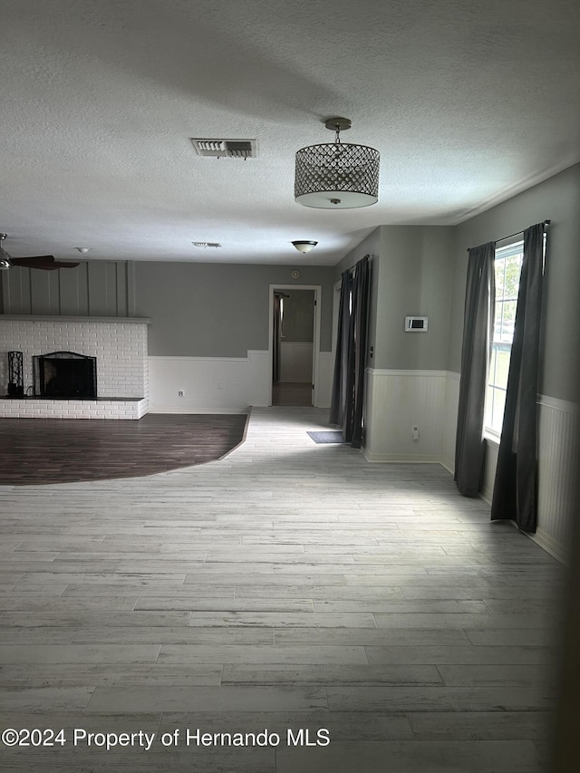 unfurnished living room featuring ceiling fan, light wood-type flooring, a textured ceiling, and a brick fireplace