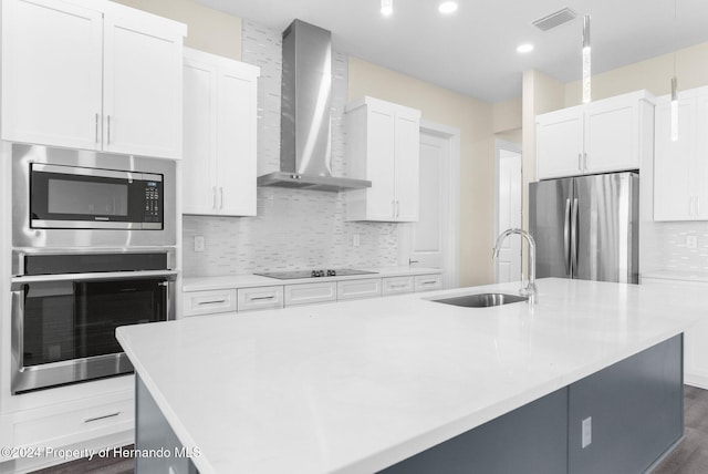 kitchen featuring stainless steel appliances, sink, wall chimney range hood, white cabinetry, and an island with sink