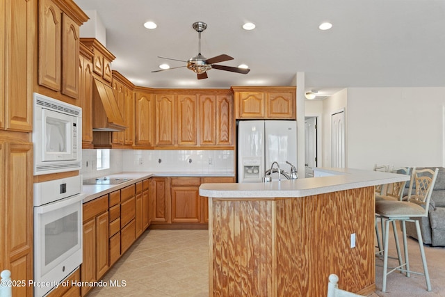 kitchen with a kitchen bar, an island with sink, white appliances, decorative backsplash, and wall chimney range hood