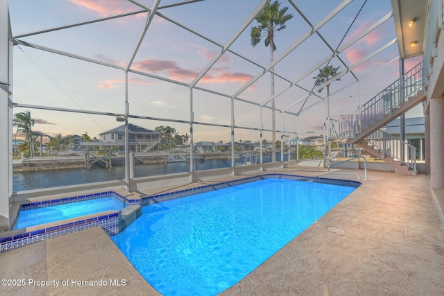 pool at dusk featuring an in ground hot tub, a water view, glass enclosure, and a patio