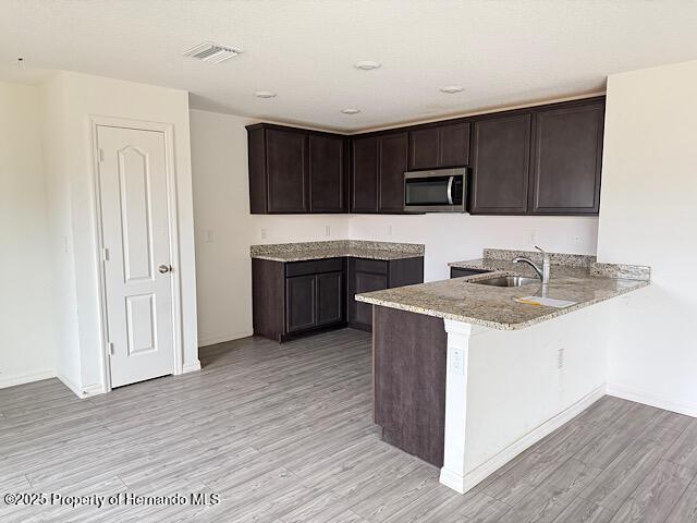 kitchen with light stone countertops, kitchen peninsula, dark brown cabinets, sink, and light hardwood / wood-style floors