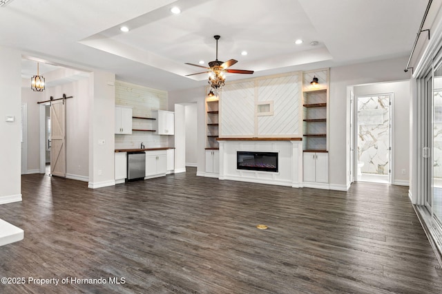 unfurnished living room with a barn door, a tray ceiling, ceiling fan, and dark wood-type flooring