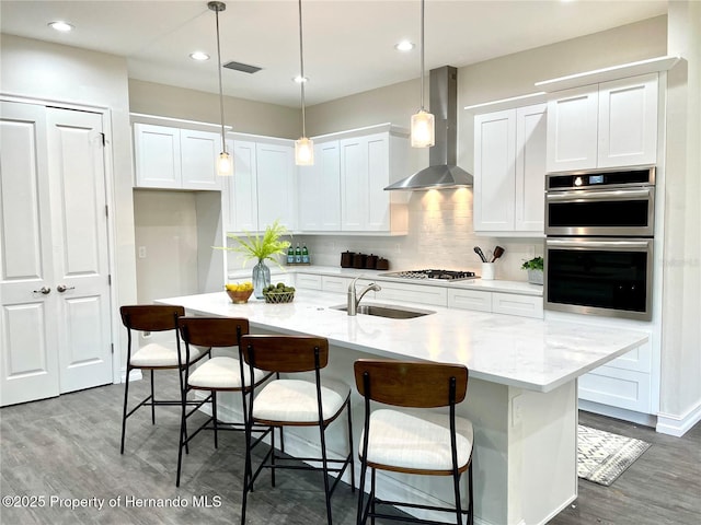 kitchen with wall chimney exhaust hood, pendant lighting, an island with sink, stainless steel appliances, and sink