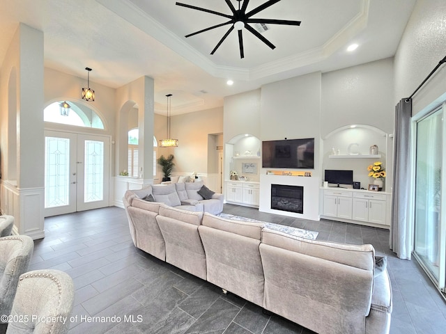 living room featuring french doors, built in features, crown molding, a tray ceiling, and ceiling fan with notable chandelier