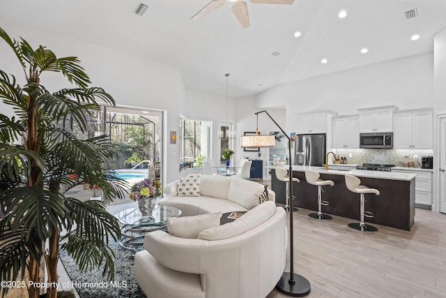 living room with ceiling fan, light wood-type flooring, sink, and high vaulted ceiling