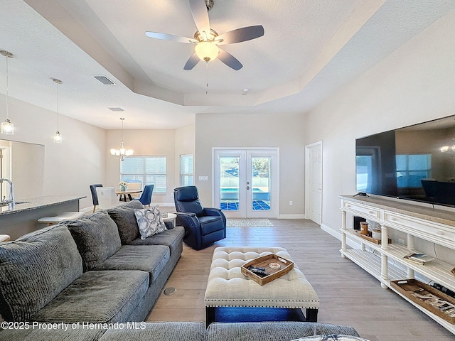 living room featuring french doors, light wood-type flooring, ceiling fan with notable chandelier, a raised ceiling, and sink