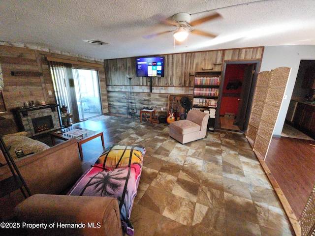 living room featuring ceiling fan, a stone fireplace, a textured ceiling, and wooden walls