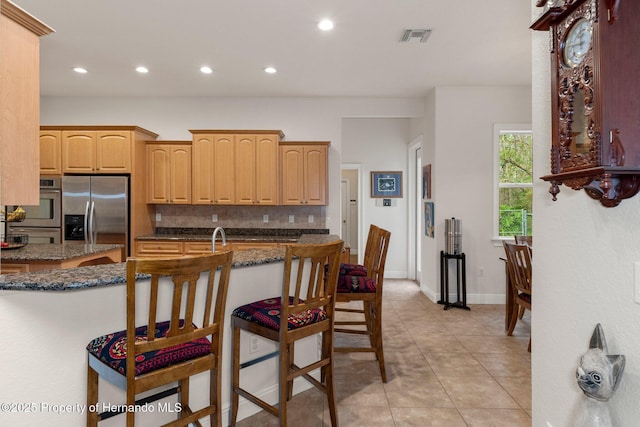 kitchen with light brown cabinets, stainless steel appliances, dark stone counters, decorative backsplash, and a breakfast bar