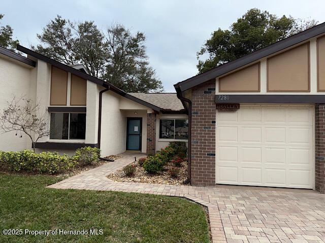 entrance to property featuring brick siding, a lawn, decorative driveway, and stucco siding