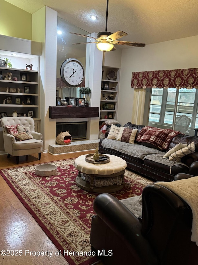 living room with built in shelves, ceiling fan, a textured ceiling, wood finished floors, and a tile fireplace