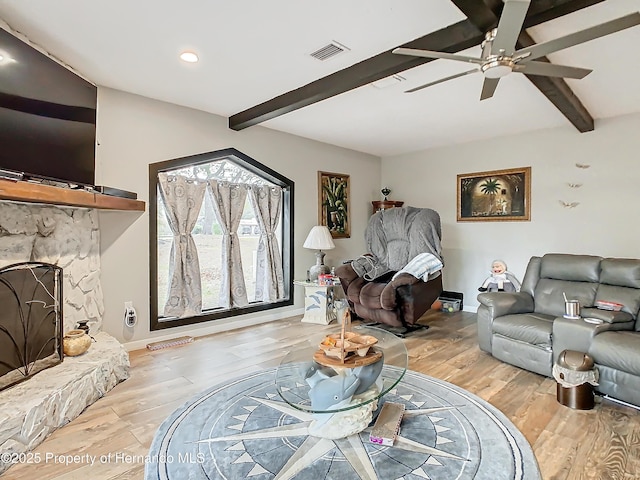 living room with ceiling fan, beamed ceiling, light hardwood / wood-style flooring, and a stone fireplace