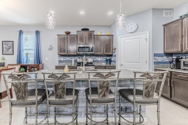 kitchen featuring dark brown cabinets, hanging light fixtures, an island with sink, stainless steel appliances, and decorative backsplash