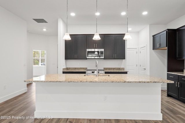 kitchen with an island with sink, dark wood-type flooring, sink, and light stone counters